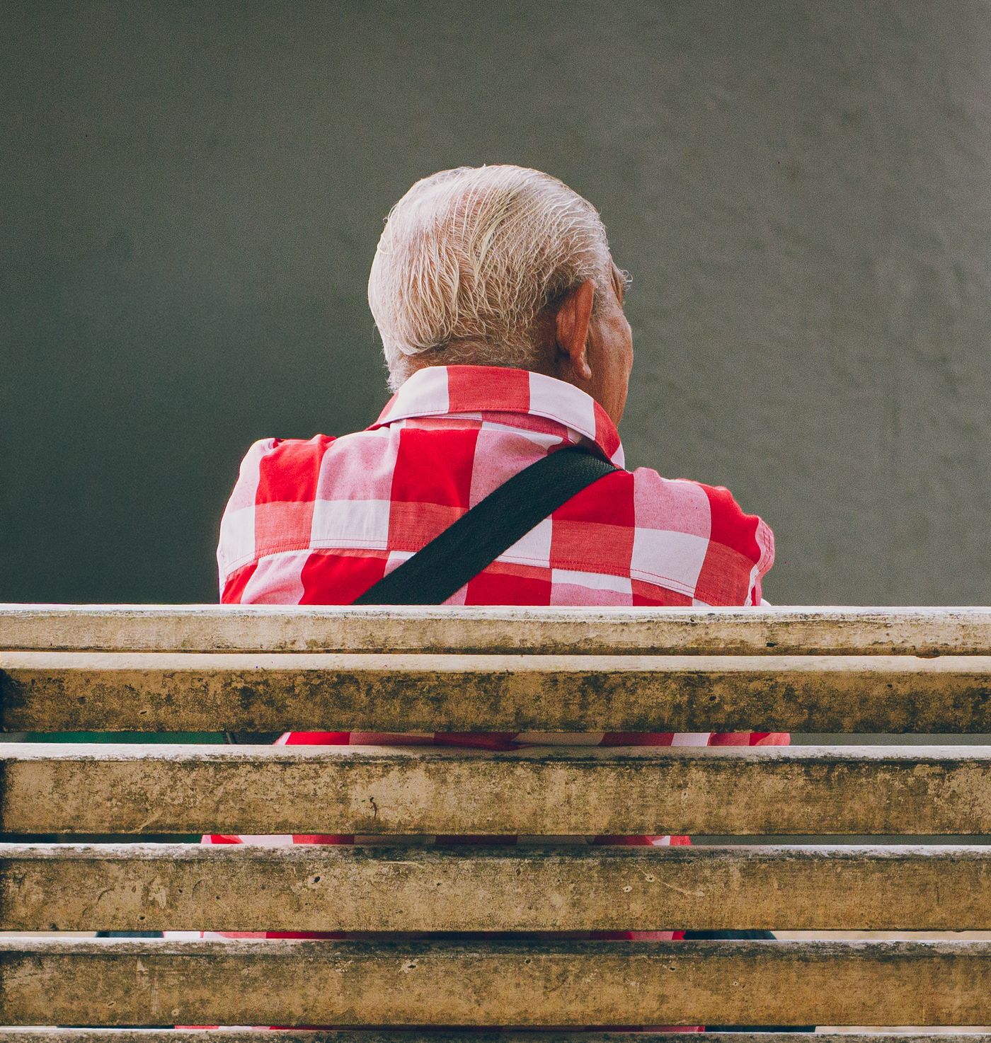 An elderly man in a red plaid shirt sitting on a bench, seen from behind