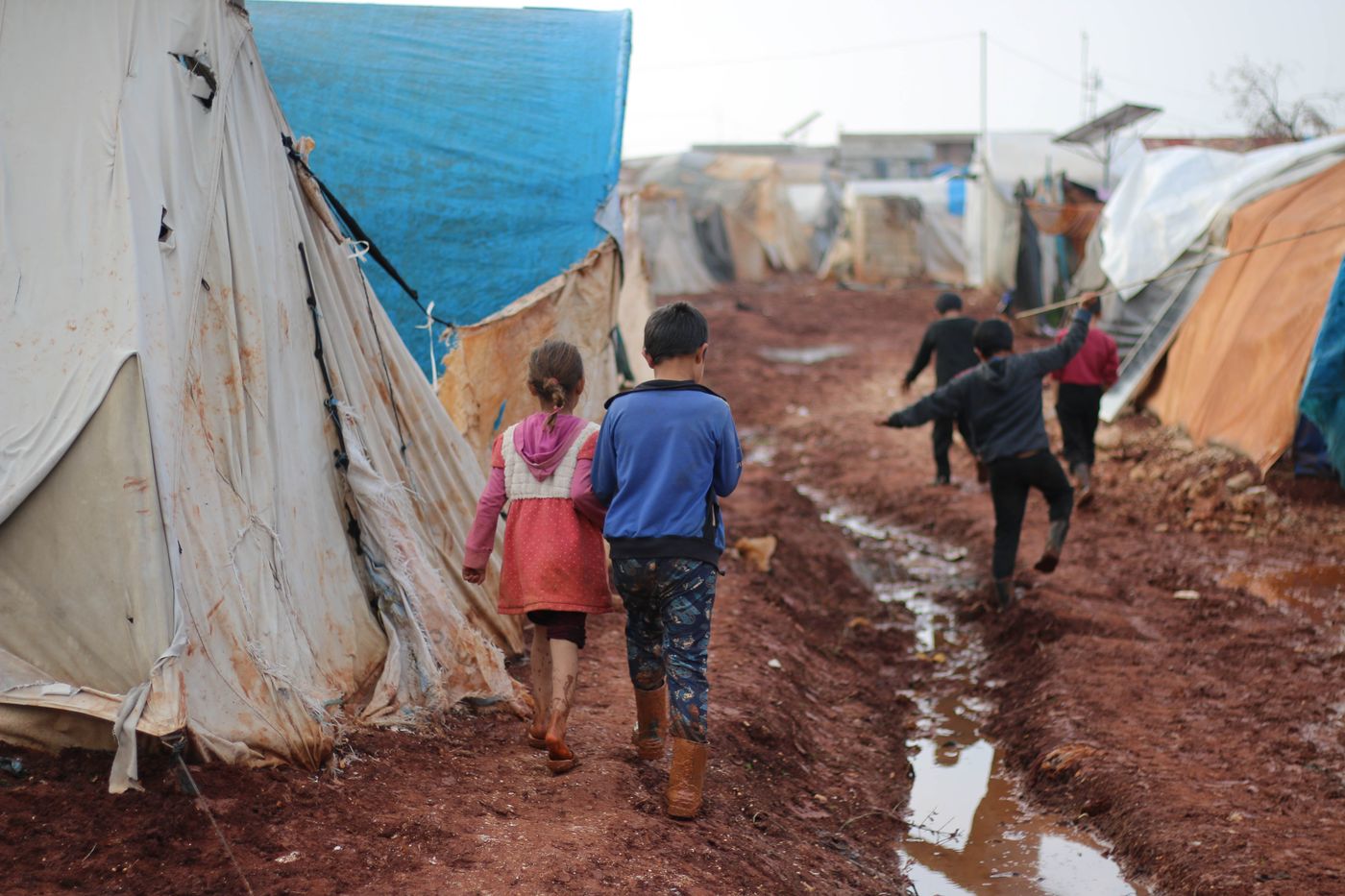Children walking down a muddy street between tents in a displacement camp, seen from behind